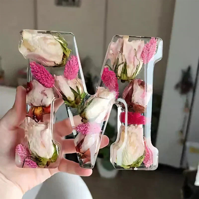 Hånd holder gennemsigtig harpiksbogstav "M" med indlejrede tørrede blomster, herunder lyserøde roser og lilla blomster. Dekorativt kunsthåndværk.