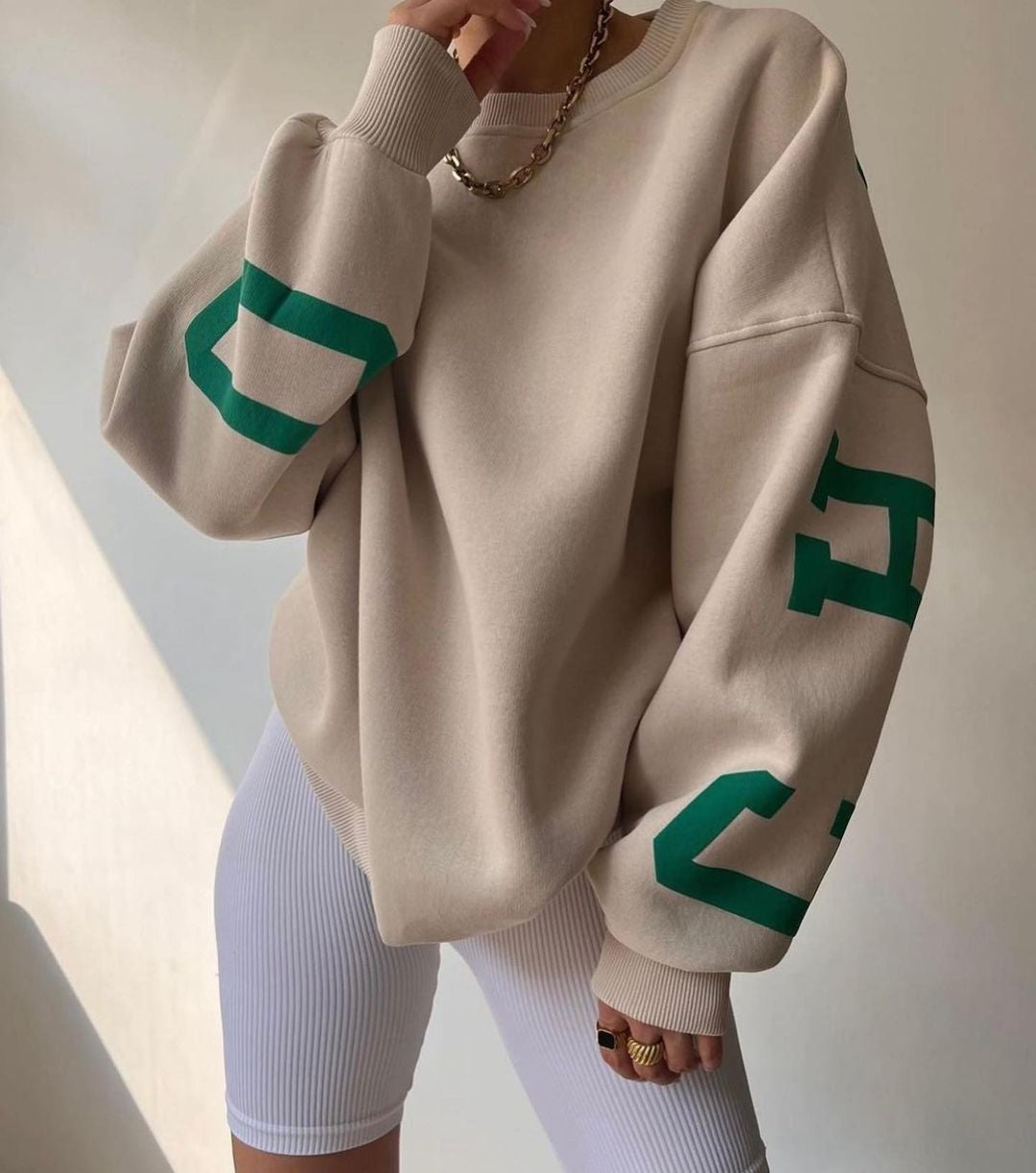 Beige oversized sweatshirt med grønne bogstaver, båret af person i hvide cykelshorts. Trendy streetwear, afslappet stil, mode 2023.