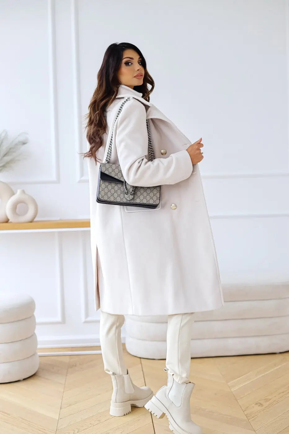 Kvinde i elegant hvid frakke med sort håndtaske, poserer i moderne stue. Mode, stilfuldt vintertøj, trendy accessories, indendørs.