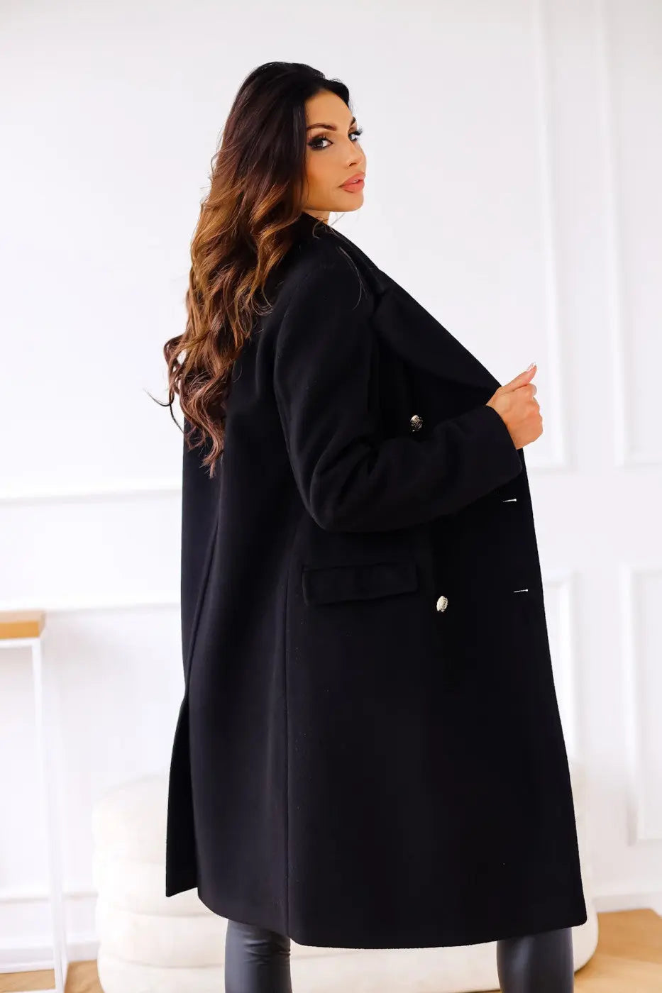 Kvinde iført elegant sort frakke med knapper, poserer i moderne interiør. Stilfuldt vintertøj, mode, damefrakke, luksusbeklædning.