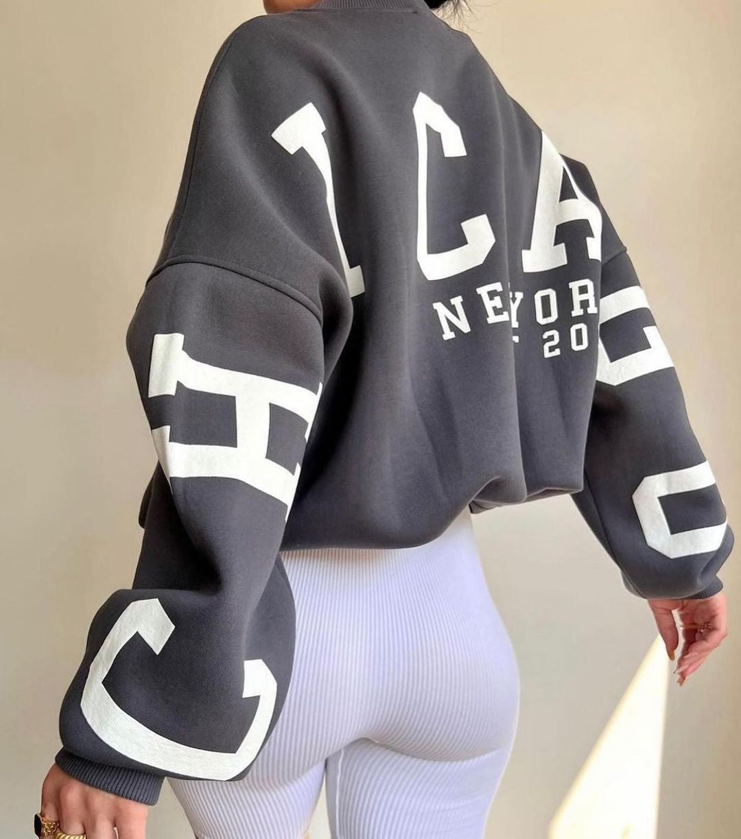 Person iført en sort sweatshirt med "Chicago New York 2020" tryk, kombineret med hvide ribbede leggings. Mode, streetwear, afslappet stil.