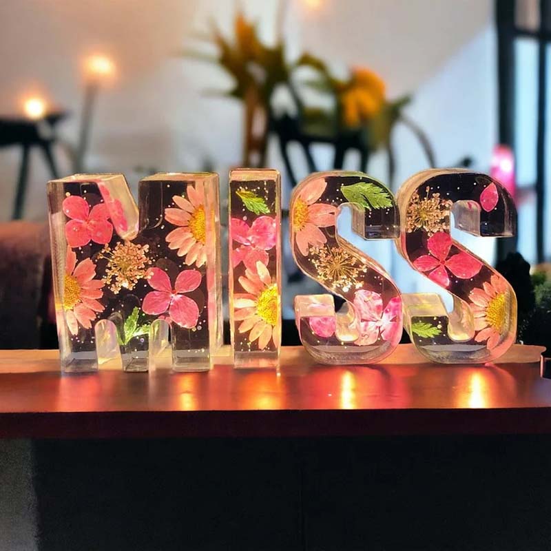 Bogstaverne "MISS" i gennemsigtig harpiks med indlejrede blomster, herunder lyserøde og gule blomster, oplyst på en hylde. Dekorativ boligindretning.