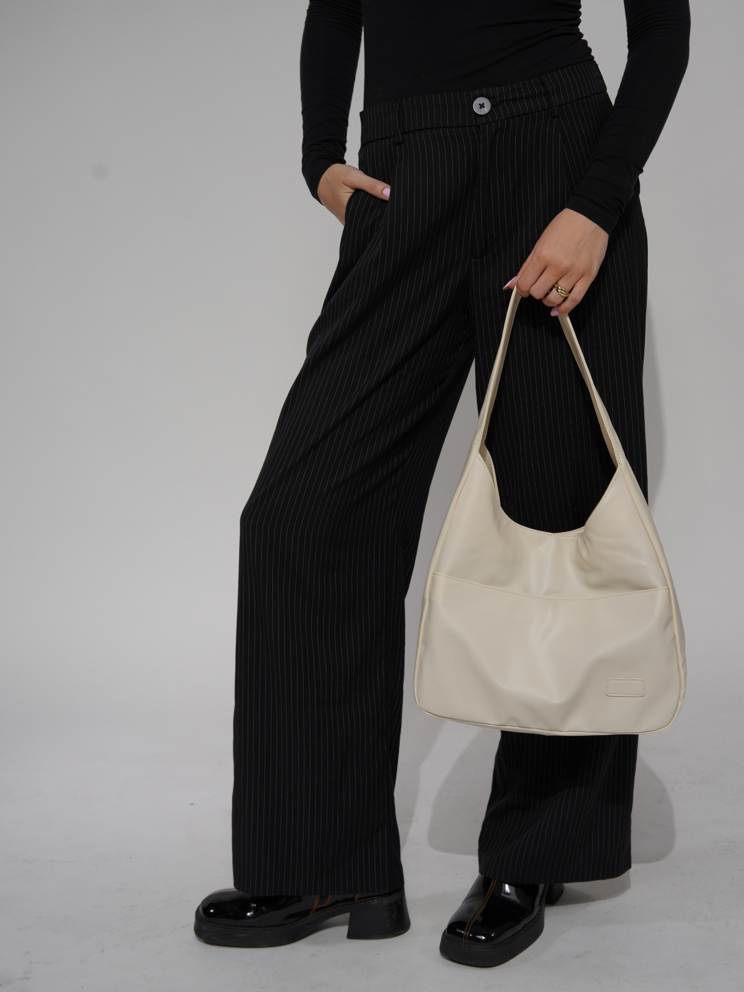 Kvinde i sorte, stribede bukser og sort trøje holder en cremefarvet skuldertaske. Mode, stilfuldt outfit, moderne tilbehør.
