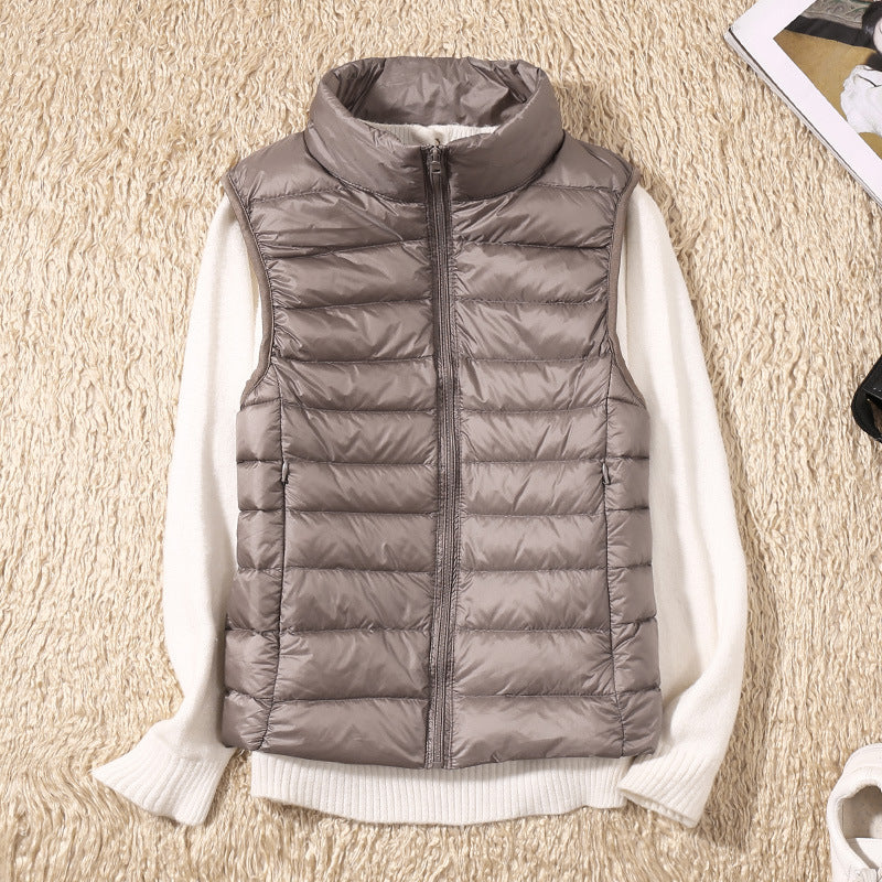 Grå dunvest med lynlås på hvid sweater, liggende på beige tæppe. Mode, vintertøj, stilfuld beklædning, varm vest, kvindemode.