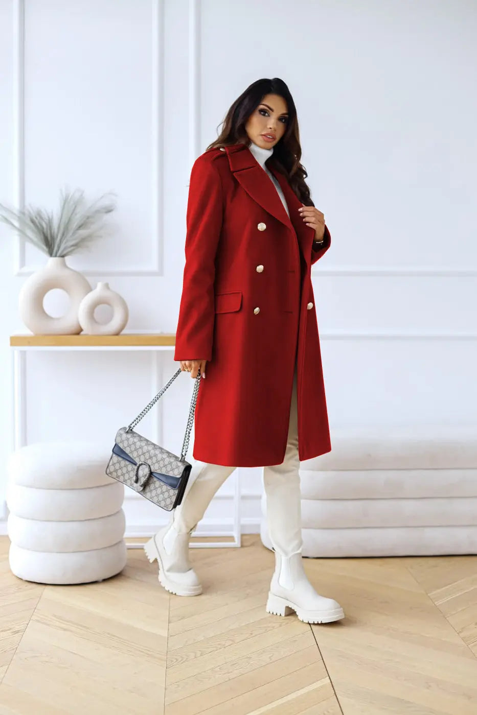 Kvinde i rød frakke med hvide støvler og designerhåndtaske, står i moderne indretning. Elegant vintermode, stilfuldt outfit, trendy accessories.