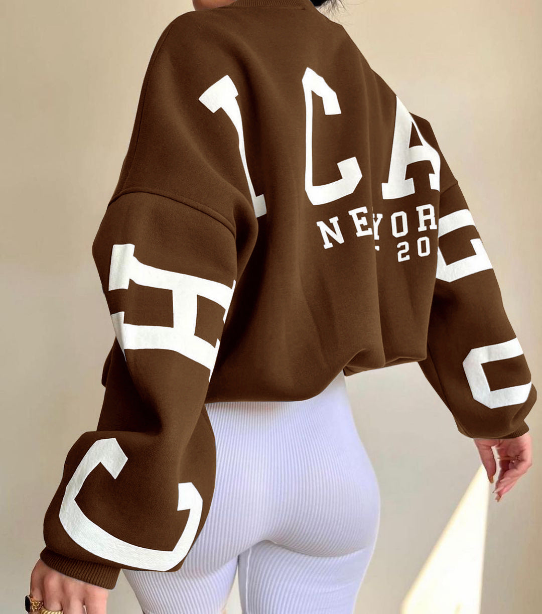 Brun sweatshirt med "Chicago New York 2023" tryk, båret af person i hvide leggings. Mode, streetwear, trendy tøj, afslappet stil.