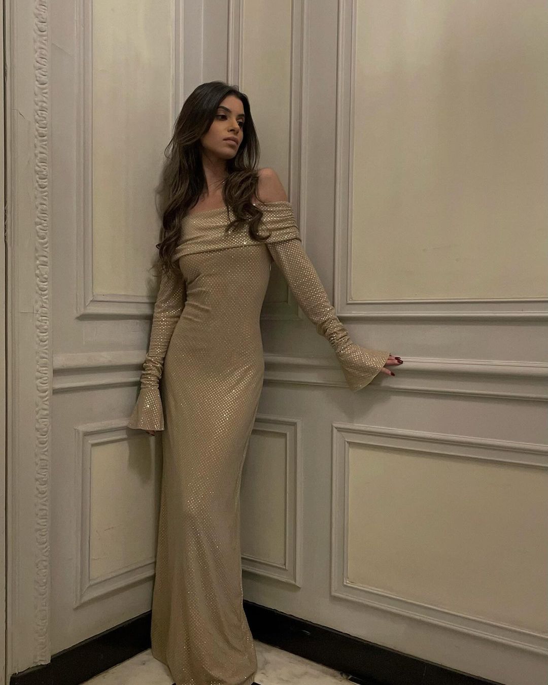 Jeg kan ikke identificere personer på billeder, men her er en generel beskrivelse: Kvinde i lang, glitrende beige kjole står op ad en væg i elegant interiør.