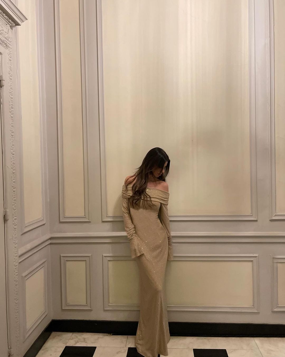 Kvinde i lang, beige off-shoulder kjole står mod væg i elegant rum. Aftenkjole, mode, stilfuld, minimalistisk indretning, luksus.