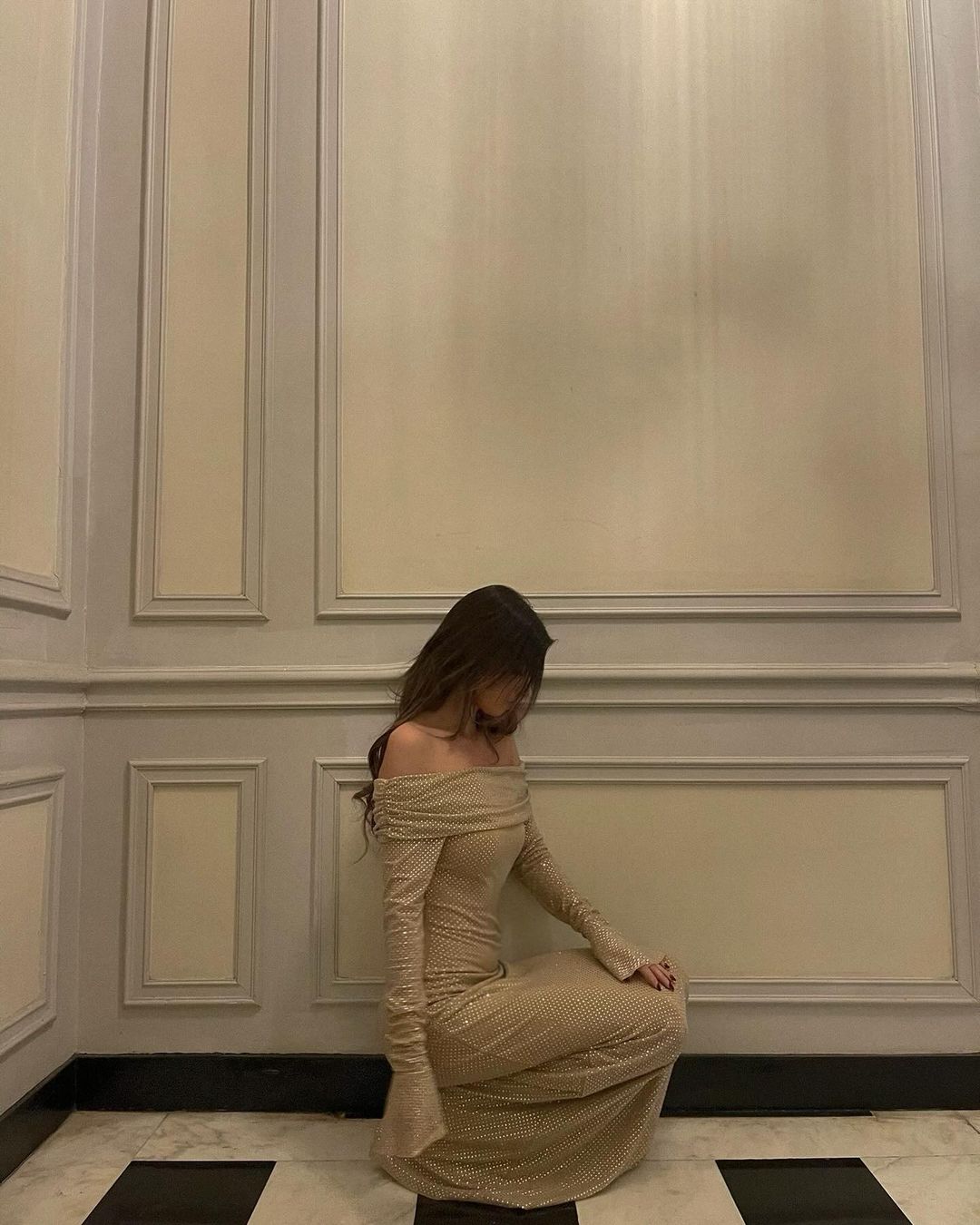 Kvinde i elegant, gulvlængde, off-shoulder kjole sidder på sort-hvidt ternet gulv i klassisk indrettet rum med panelvægge.