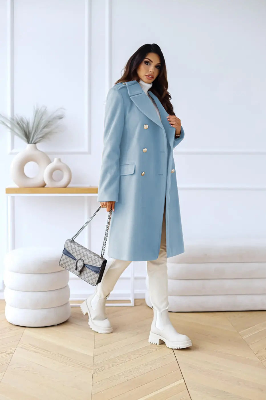 Kvinde i lyseblå frakke med guldknapper, hvidt tøj og støvler, holder designerhåndtaske. Moderne mode, elegant stil, indendørs.