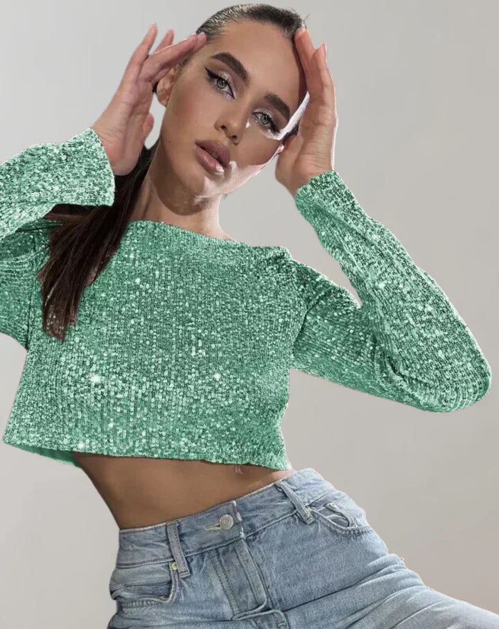Kvinde i grøn glitrende top og blå jeans, poserer med hænderne ved ansigtet. Moderne mode, glamourøst look, trendy tøjstil.