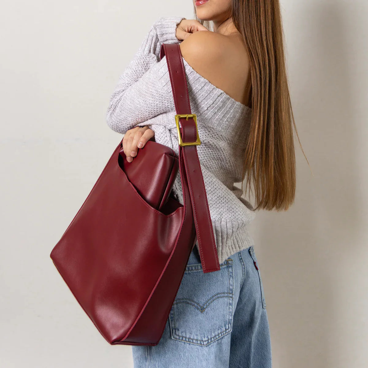 Kvinde med langt hår bærer en stor, rød lædertaske over skulderen. Hun har en grå sweater og blå jeans på. Mode, stilfuld taske, trendy tilbehør.
