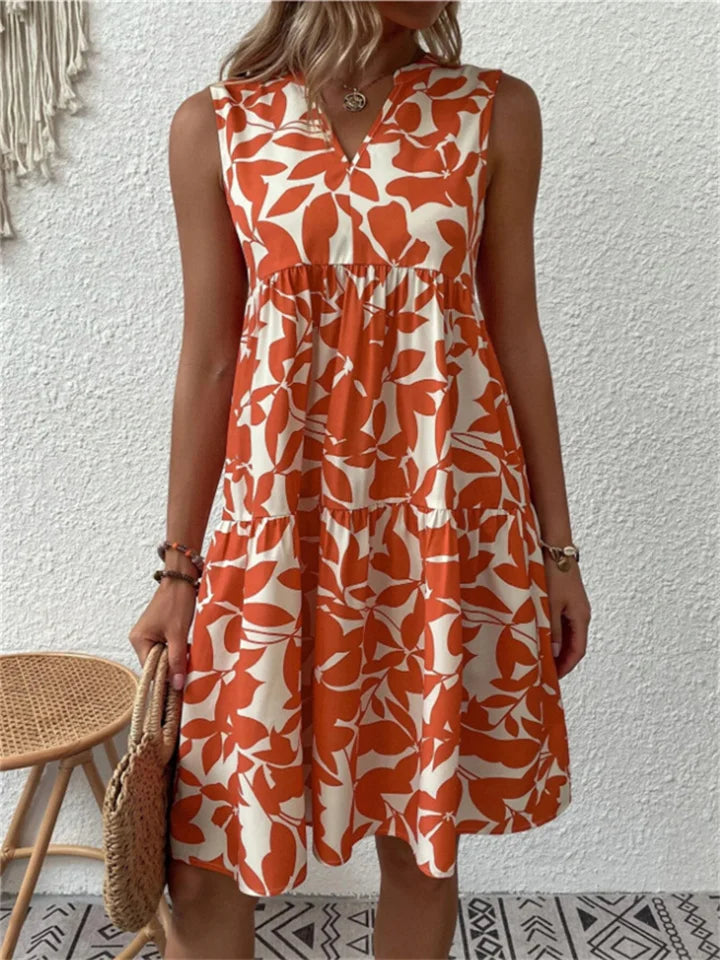 Kvinde i orange-hvid blomstret sommerkjole, står foran hvid væg. Kjolen har ærmeløst design og A-formet snit. Perfekt til sommermode.