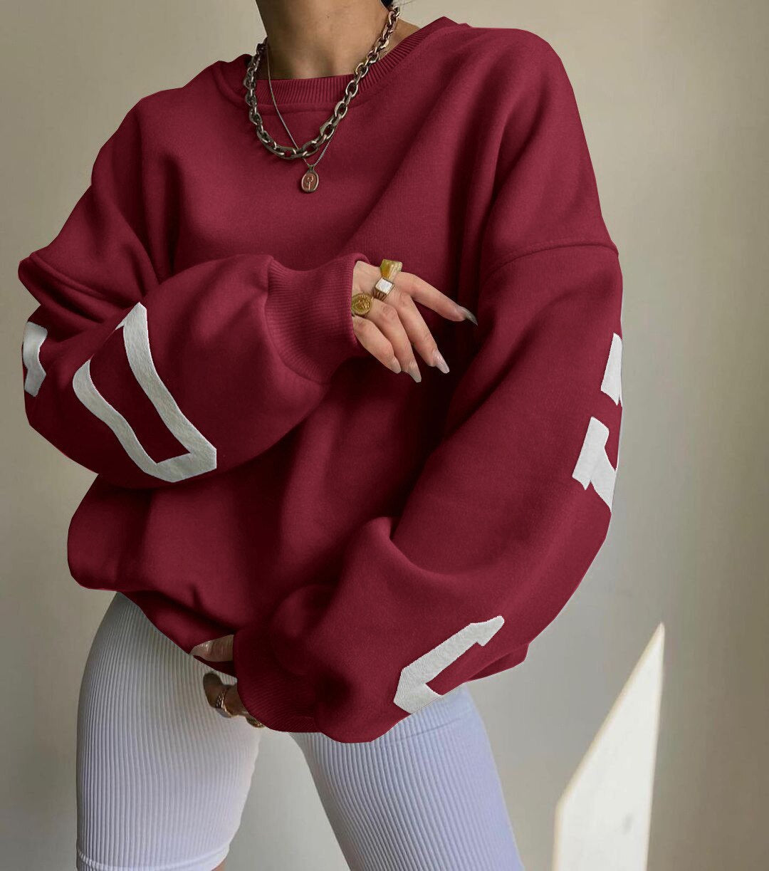 Rød oversized sweatshirt med hvide tal, båret af person med guldringe og halskæde. Moderne streetwear-stil, populær mode 2023.