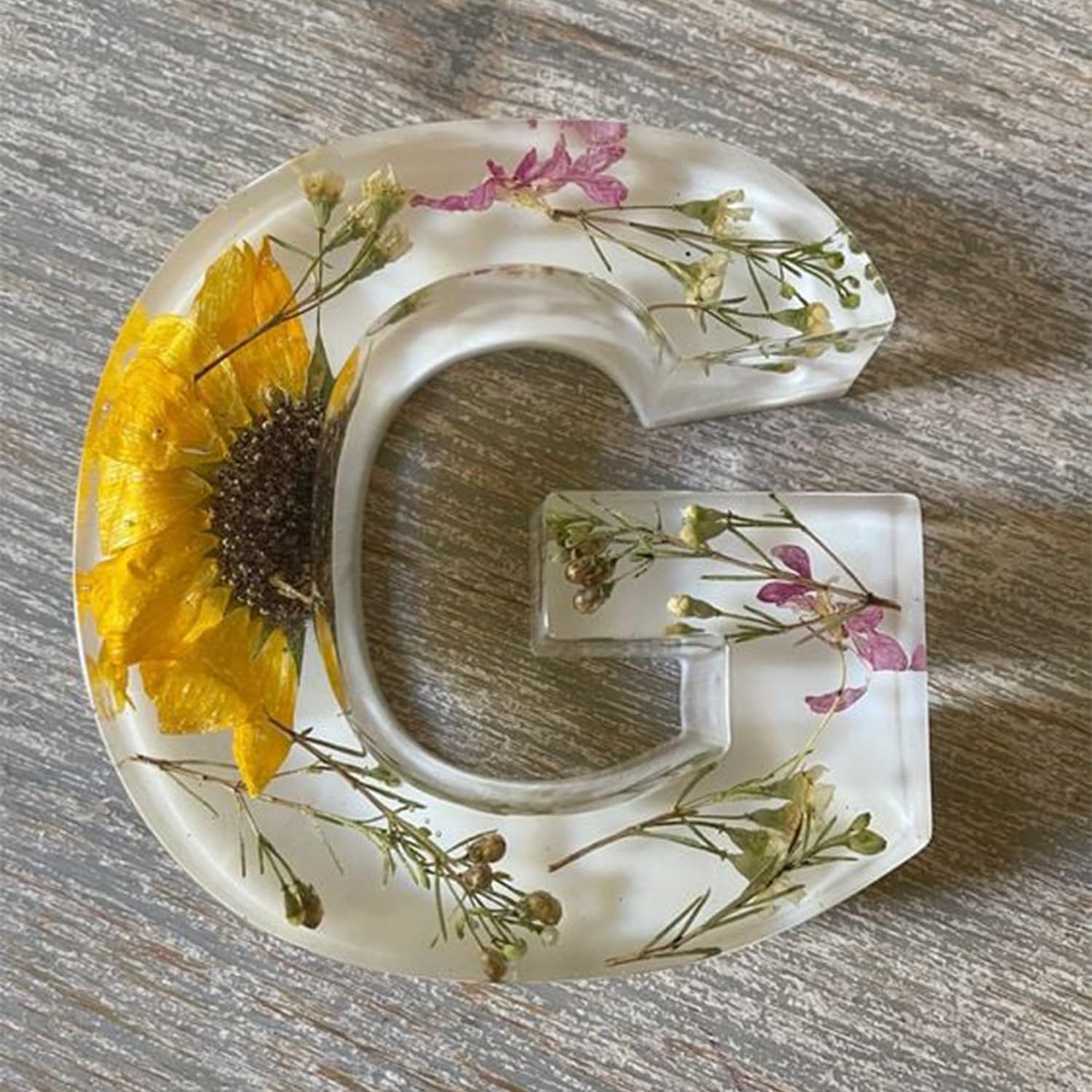 Bogstavet G i gennemsigtig harpiks med indlejrede tørrede blomster, herunder en gul solsikke, på en træoverflade. Dekorativt og unikt design.