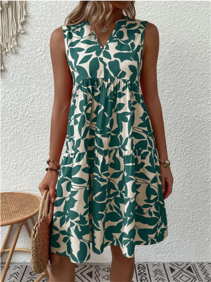 Kvinde i grøn-hvid blomstret sommerkjole, står foran hvid væg. Kjolen har ærmeløst design og A-formet snit. Perfekt til sommermode og casual stil.