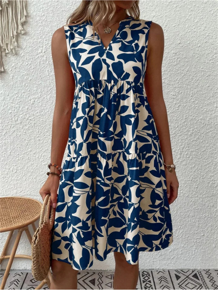 Kvinde i blå-hvid blomstret sommerkjole, står foran hvid væg. Kjolen er ærmeløs med A-line snit. Perfekt til sommermode og casual stil.