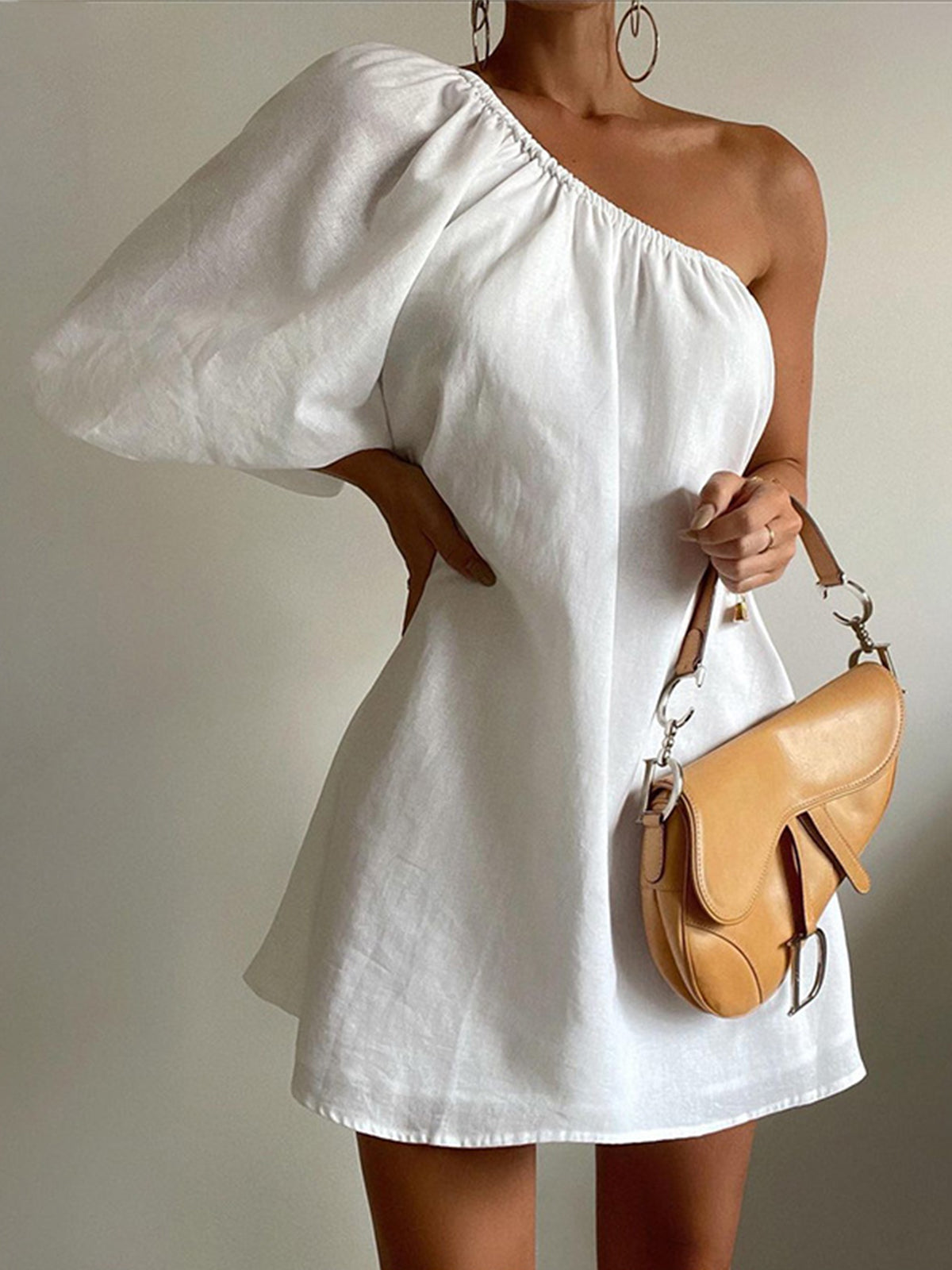 Kvinde i hvid asymmetrisk kjole med én skulder, holder en beige lædertaske. Elegant sommermode, minimalistisk stil, trendy accessories.