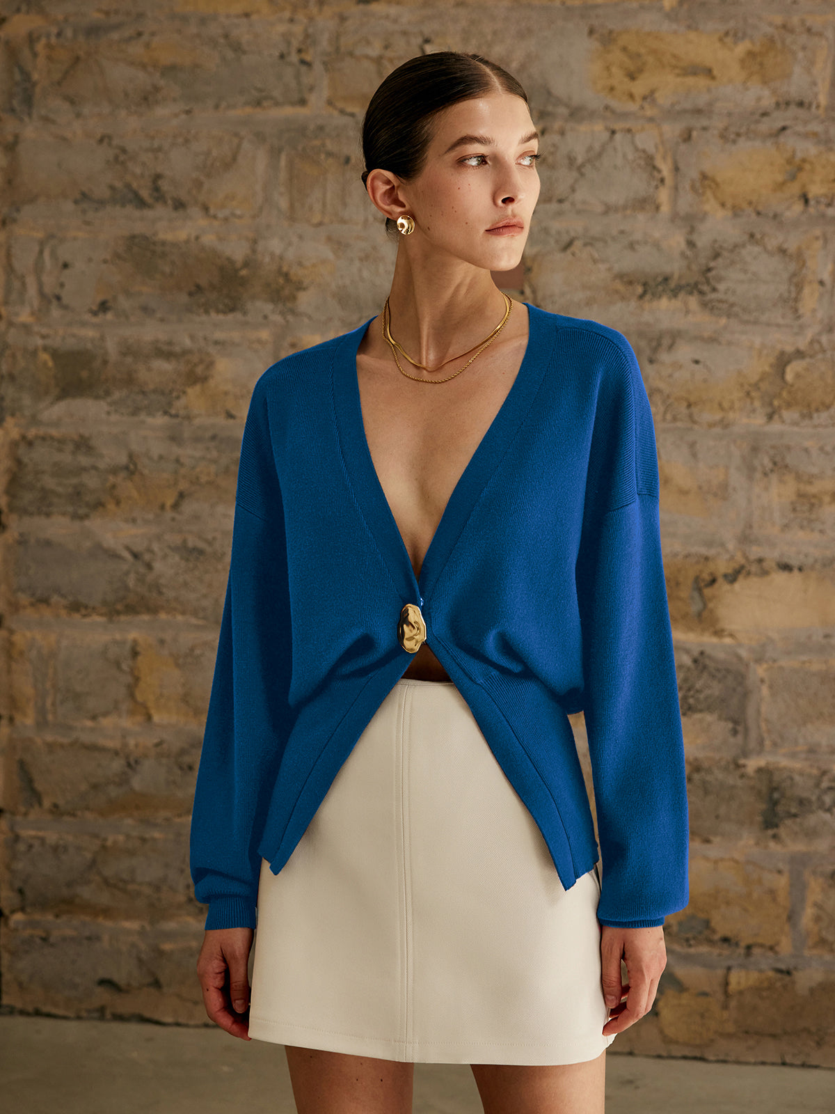 Kvinde iført blå cardigan med guldknap og hvid nederdel foran murstensvæg. Elegant mode, minimalistisk stil, moderne kvindetøj.
