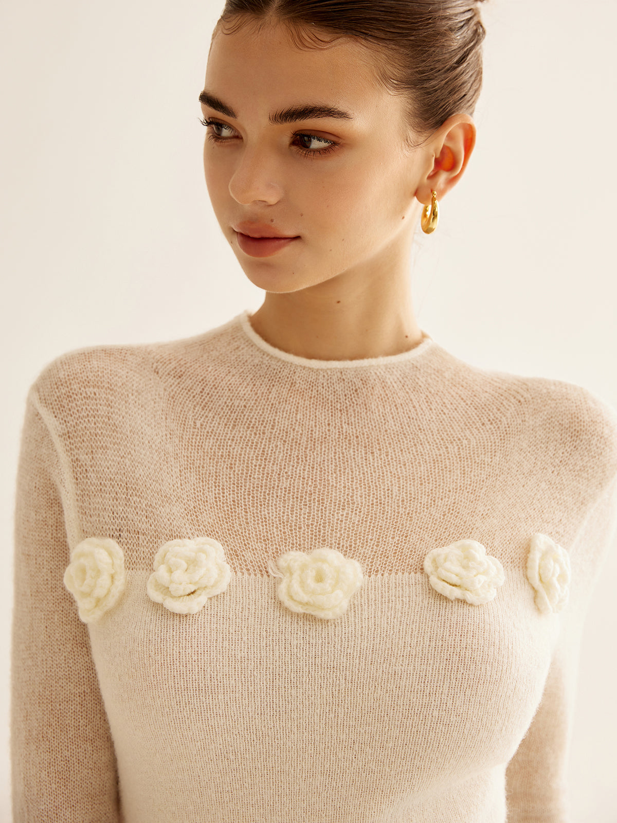Kvinde iført beige striktrøje med blomsterdetaljer, gyldne øreringe. Elegant mode, minimalistisk design, trendy tøjstil.