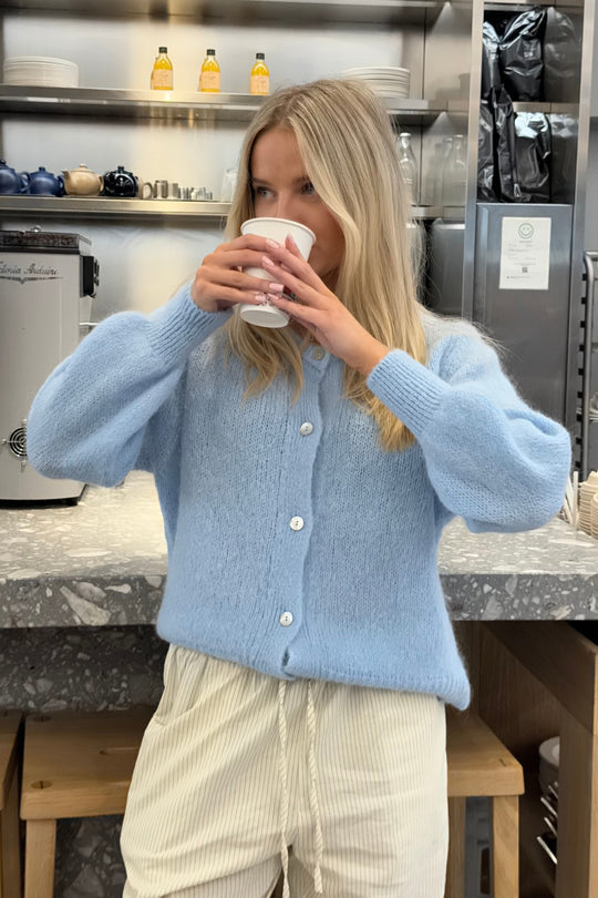 Kvinde i lyseblå cardigan drikker kaffe i moderne café. Baggrund med kaffemaskine og hylder med kopper. Hygge, café, mode, afslapning.
