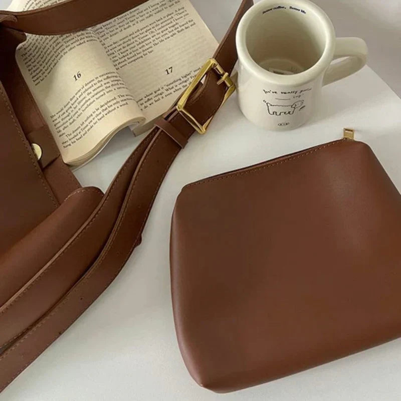 Brun lædertaske og pung ved siden af en åben bog og en hvid kaffekop på et bord. Stilfuldt tilbehør, mode, lædervarer, minimalistisk design.