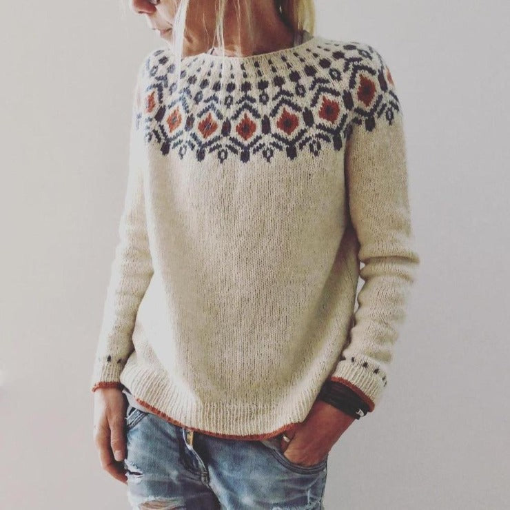 Kvinde iført beige striktrøje med nordisk mønster og blå jeans. Trendy vintermode, skandinavisk design, casual stil.