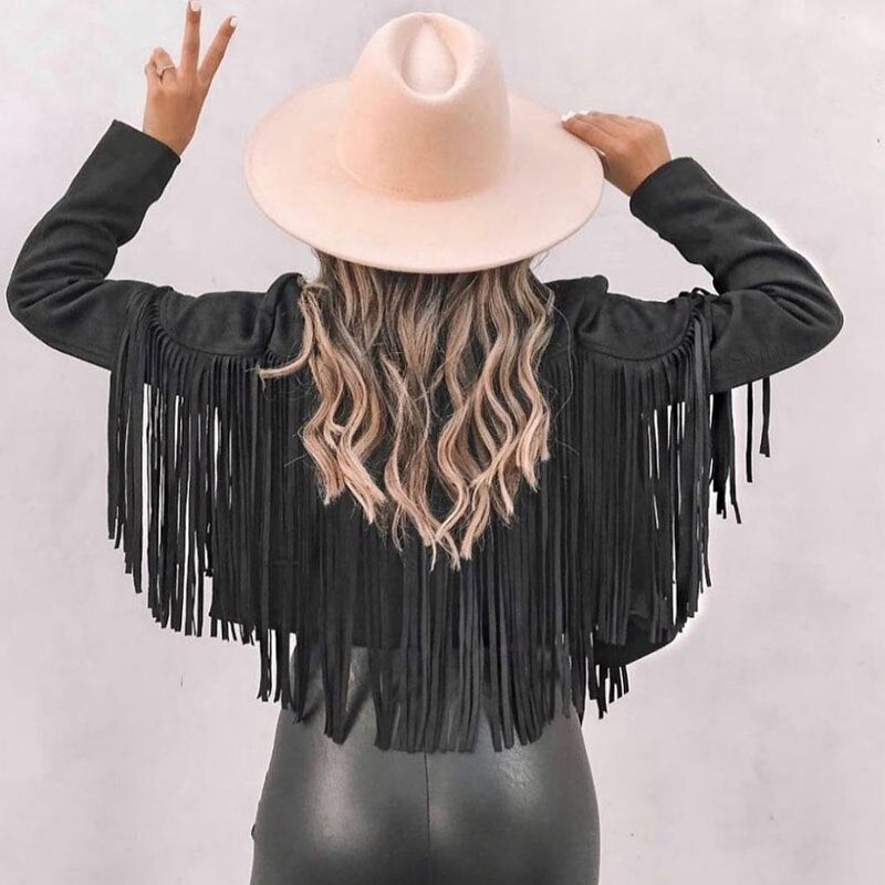 Kvinde med sort frynsejakke og beige hat, set bagfra. Hun har bølget hår og viser fredstegn. Mode, boho-stil, trendy outfit.