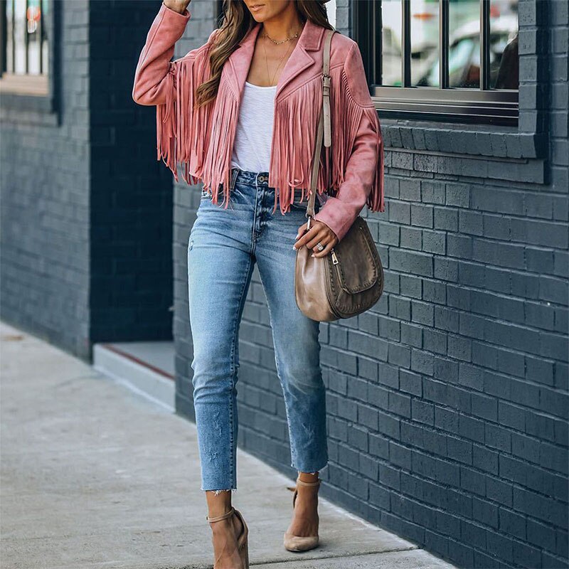 Kvinde iført lyserød frynsejakke, hvid top, blå jeans og beige hæle, bærer brun taske. Mode, street style, trendy outfit, bymiljø.