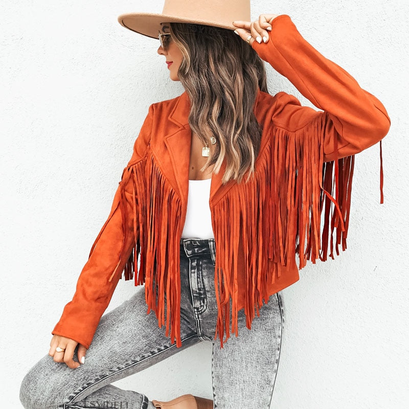 Kvinde i orange frynsejakke og bredskygget hat, sidder mod hvid væg. Mode, efterårstøj, casual stil, trendy jakke, streetwear look.