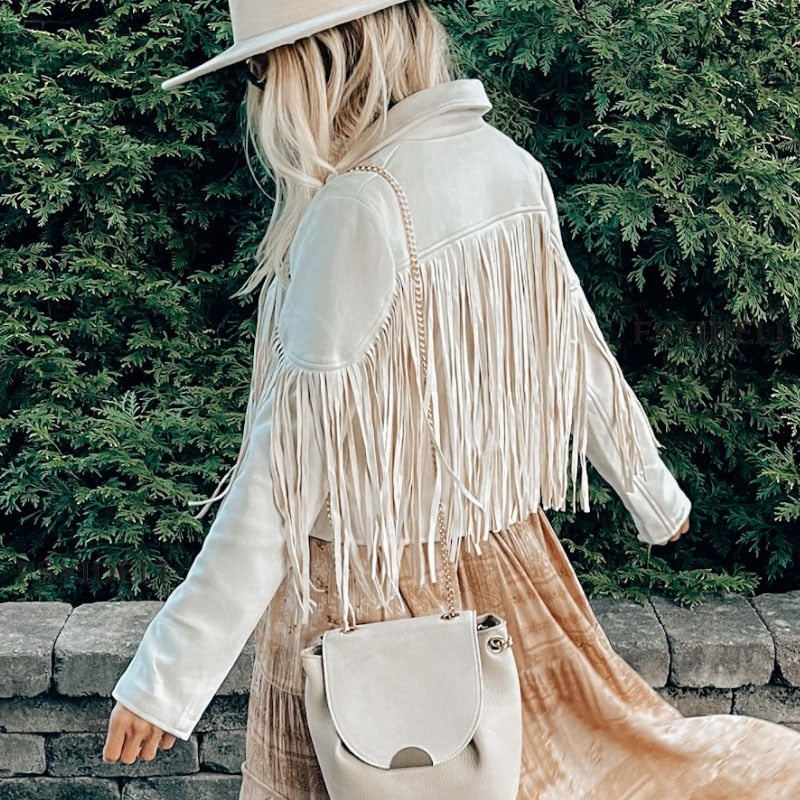 Kvinde i hvid frynsejakke og hat, bærer beige taske. Står foran grøn busk. Mode, stil, efterårstøj, trendy jakke, kvindemode.