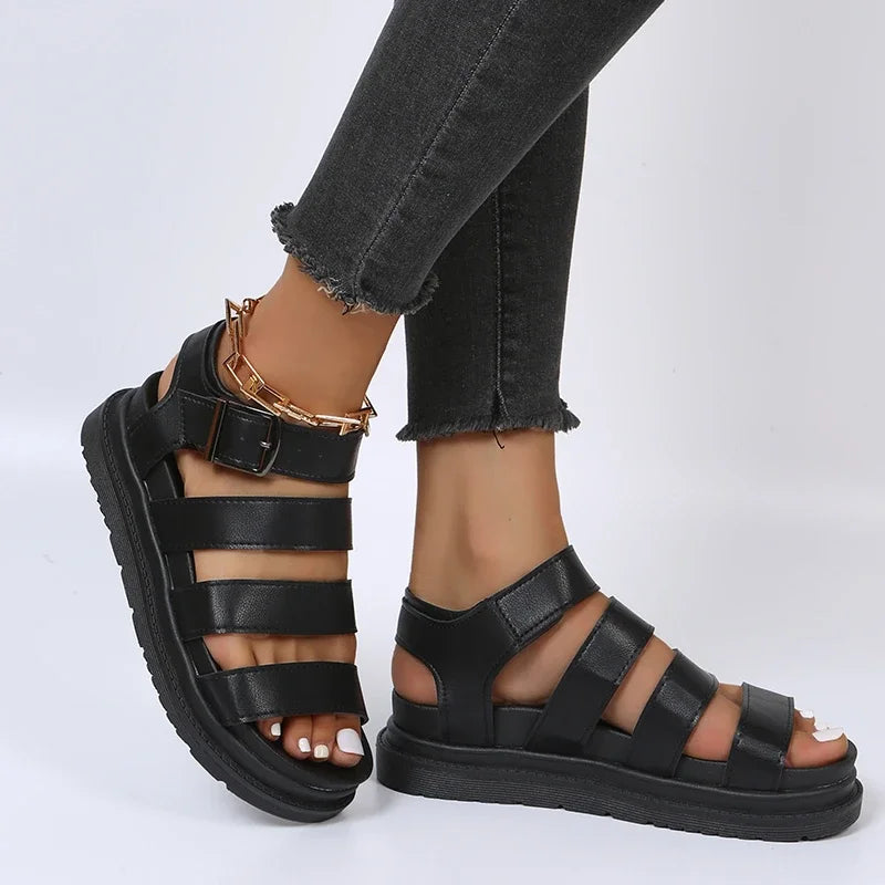 Sort læder sandaler med remme og guldspænde, båret af person i sorte jeans. Komfortable, moderne fodtøj til sommermode og casual stil.