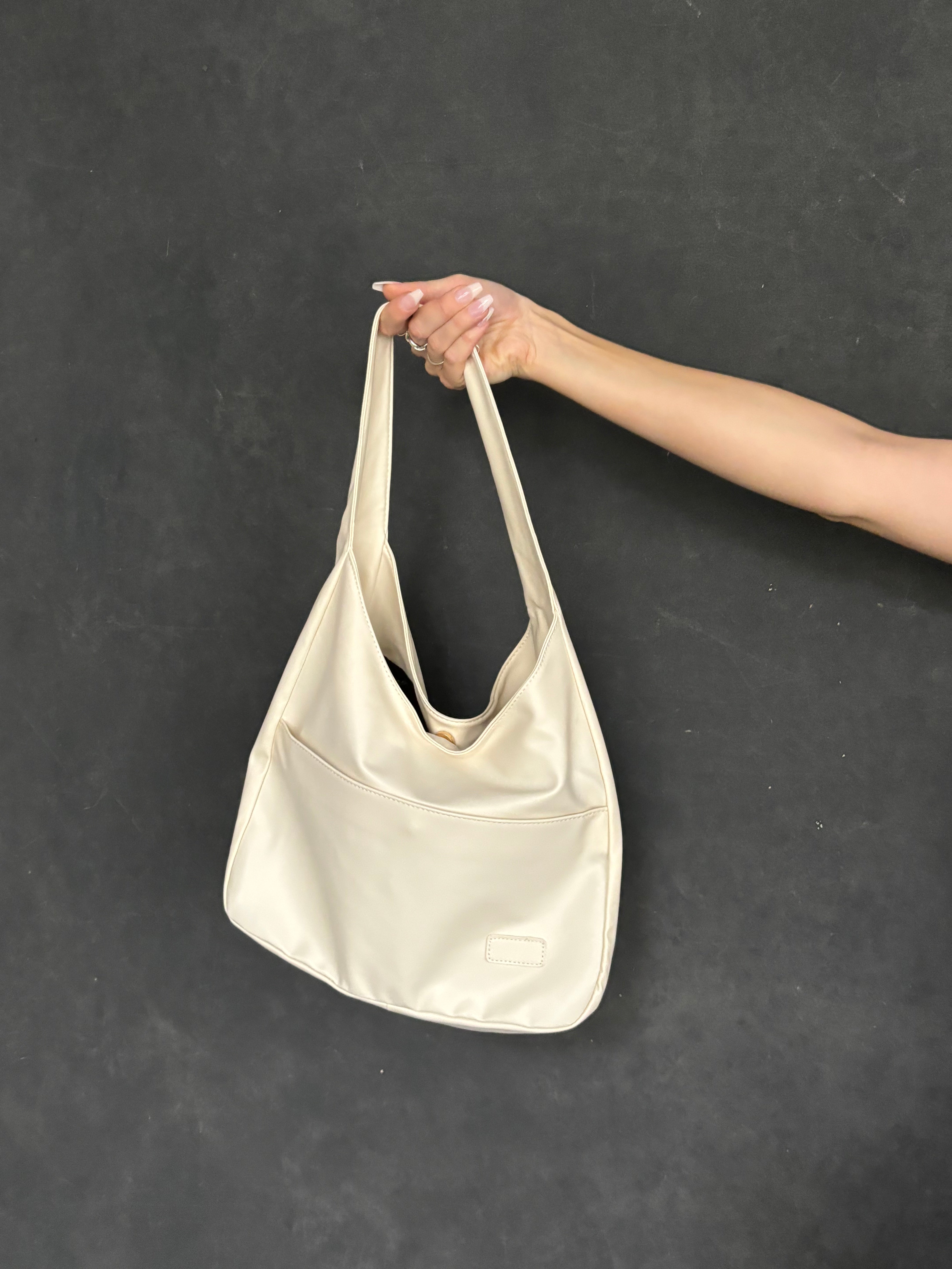 Beige skuldertaske i læder holdt af en hånd mod sort baggrund. Stilfuld og moderne design, perfekt til hverdagsbrug.