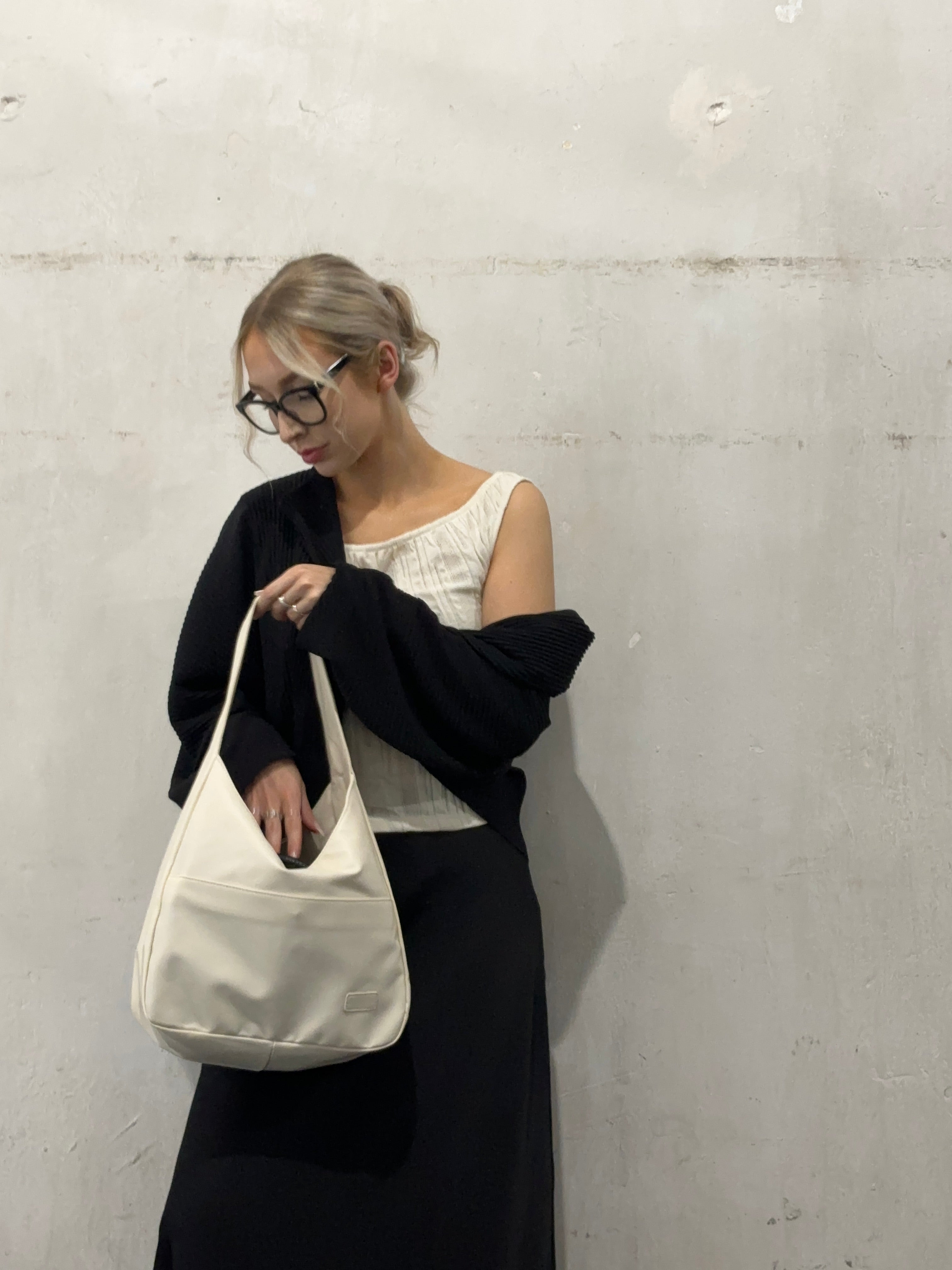 Kvinde med briller iført sort cardigan og hvid top, holder en hvid skuldertaske. Minimalistisk mode, moderne stil, betonvæg baggrund.