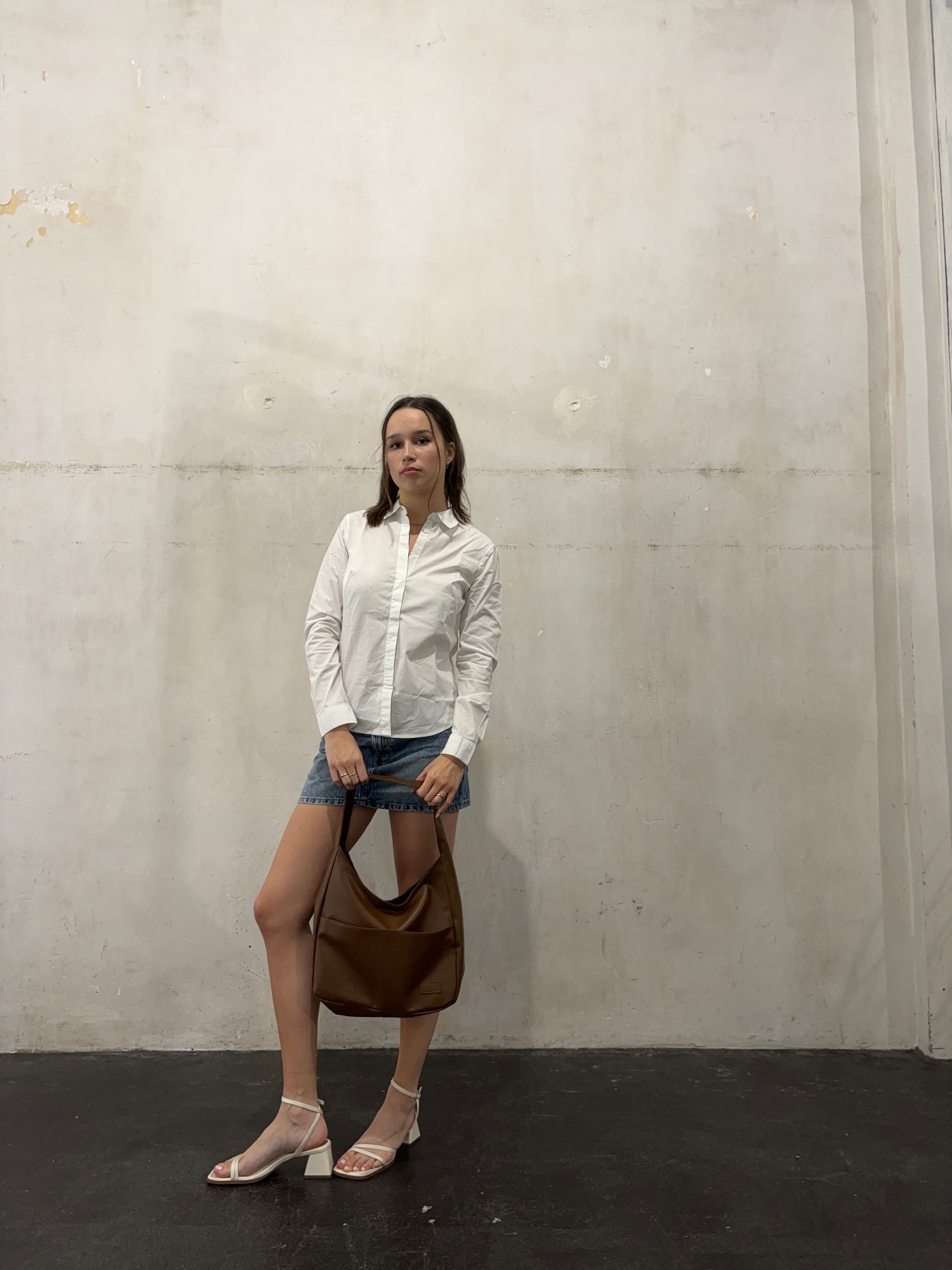 Ung kvinde i hvid skjorte og denimnederdel står mod en rå betonvæg, holder en brun lædertaske. Mode, minimalistisk stil, casual outfit.