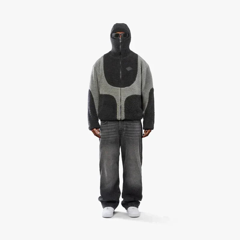 Person iført grå og sort hættetrøje med lynlås, hætte og solbriller, grå jeans og hvide sneakers. Moderne streetwear mode.