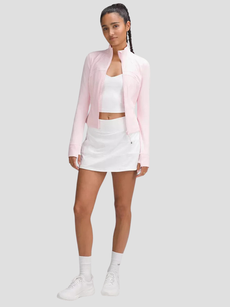 Kvinde iført lyserød jakke, hvid top og hvid nederdel. Sporty outfit, moderigtigt tøj, aktiv livsstil, moderne kvindemode, komfortabelt design.