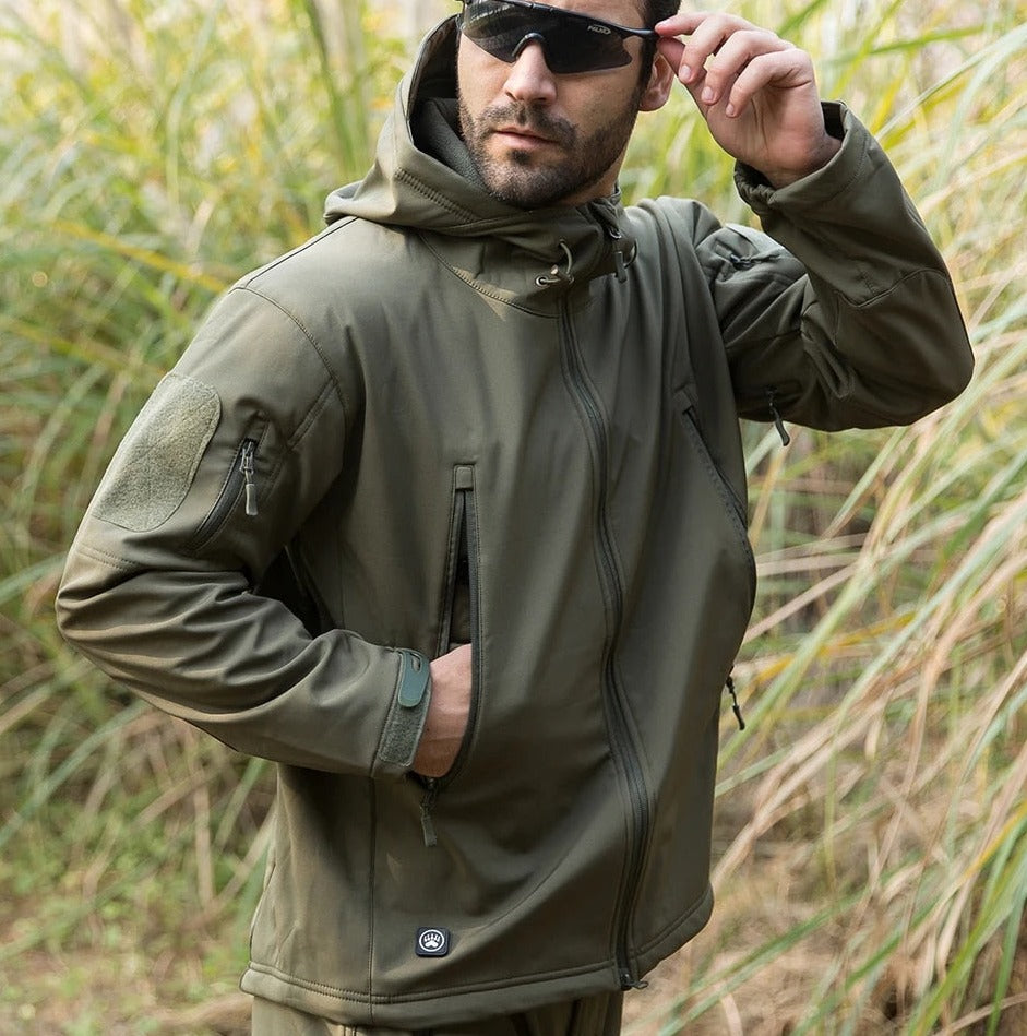 Mand iført olivengrøn softshell-jakke med hætte og solbriller i naturmiljø. Taktisk jakke, udendørs beklædning, camouflage, friluftsliv.