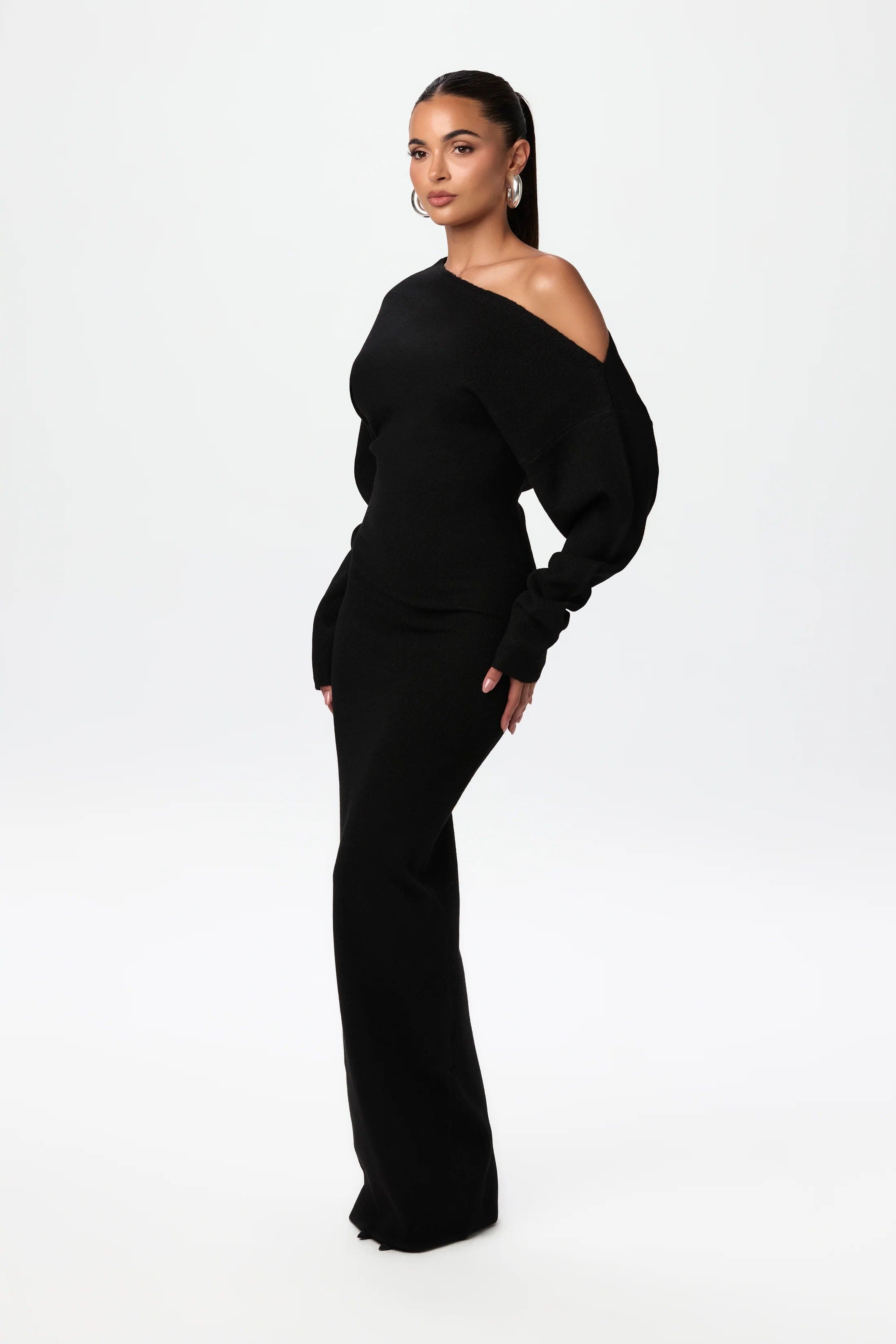 Kvinde i elegant sort off-shoulder kjole, lange ærmer, poserer mod hvid baggrund. Stilfuldt modebillede, perfekt til aftenanledninger.