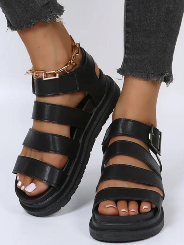 Sort læder sandaler med remme og spænde, båret af person med ankelsmykke og hvide negle. Perfekt til sommermode og casual stil.