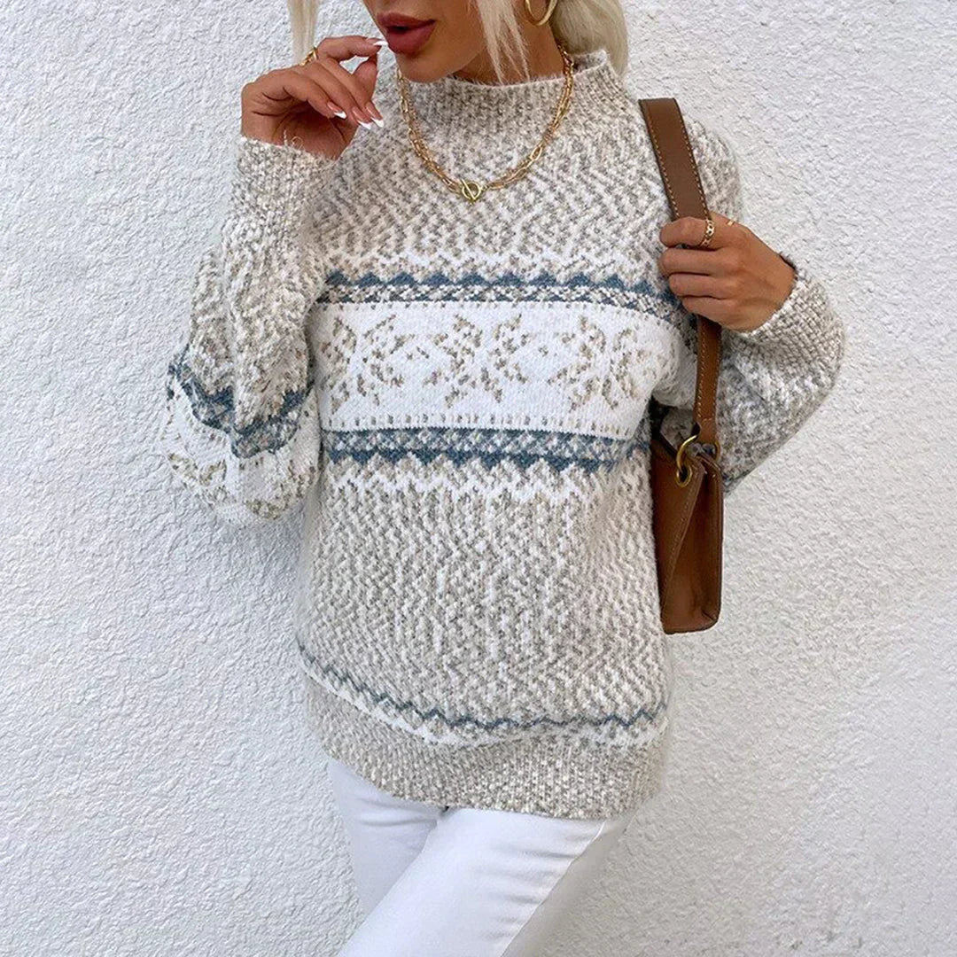 Kvinde iført strikket sweater med nordisk mønster, beige og blå nuancer, accessoriseret med guldhalskæde og brun taske, mod hvid væg.