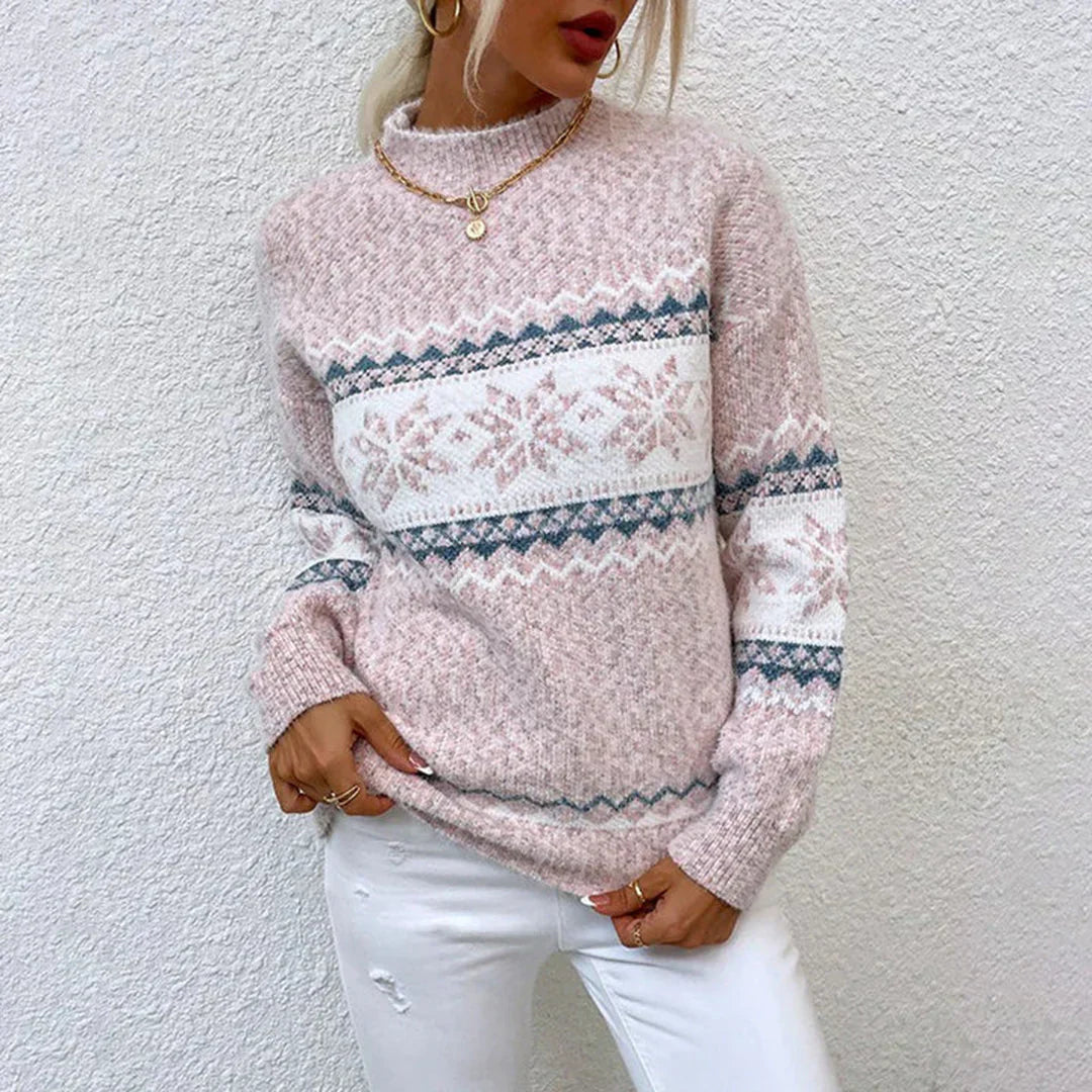 Kvinde iført lyserød striktrøje med snefnugmønster, hvid baggrund, guldhalskæde, hvide jeans. Vintermode, stilfuldt tøj, trendy strik.