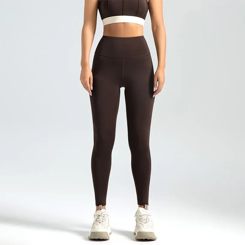Kvinde iført brune træningstights og sports-bh, stående mod en neutral baggrund. Fitnessmode, komfortabelt sportstøj, høj talje leggings.