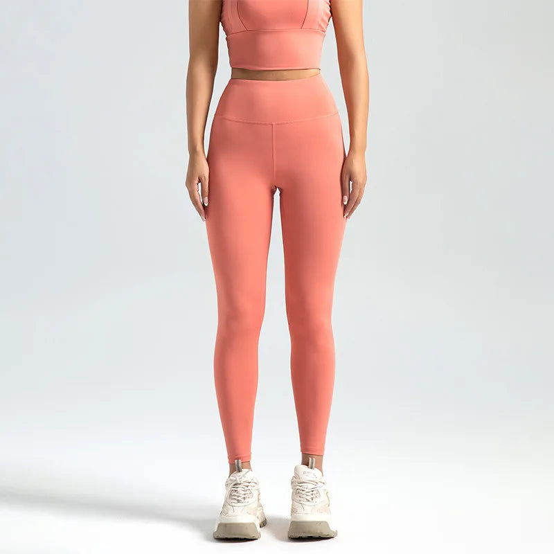 Kvinde iført lyserødt træningssæt med leggings og top, står mod neutral baggrund. Sportstøj, fitnessmode, aktiv livsstil.