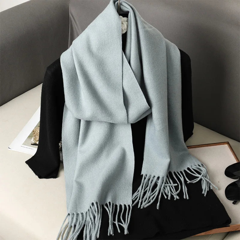 Lyseblå kashmir tørklæde med frynser på en sort sweater, placeret på en sofa. Elegant og stilfuldt tilbehør til vintermode.
