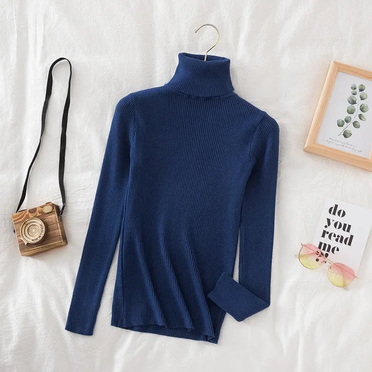 Blå rullekrave sweater på hvidt sengetøj, ved siden af retro kamera, solbriller og indrammet billede. Moderne mode, minimalistisk stil.
