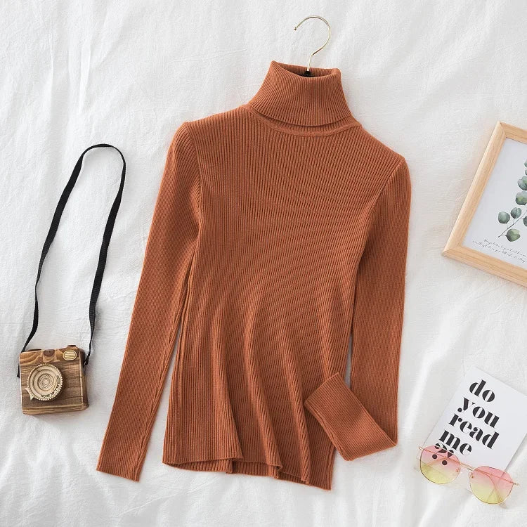 Brun rullekrave sweater i ribstrik på hvid baggrund, ved siden af et kamera, solbriller og en billedramme. Perfekt til efterårsmode og stil.