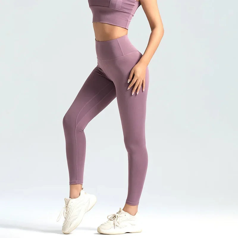 Kvinde iført lilla træningstøj, herunder sports-bh og leggings, poserer mod lys baggrund. Komfortabelt og stilfuldt fitnessoutfit med hvide sneakers.