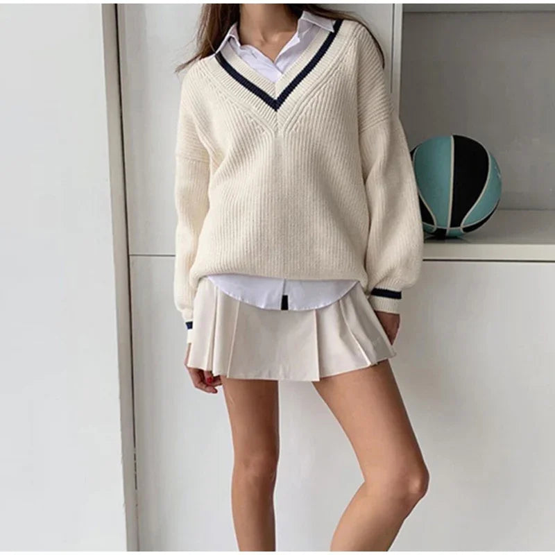 Kvinde iført hvid sweater med V-hals og plisseret nederdel, står ved hvid reol med blå og sort fodbold. Mode, efterårstøj, casual stil.