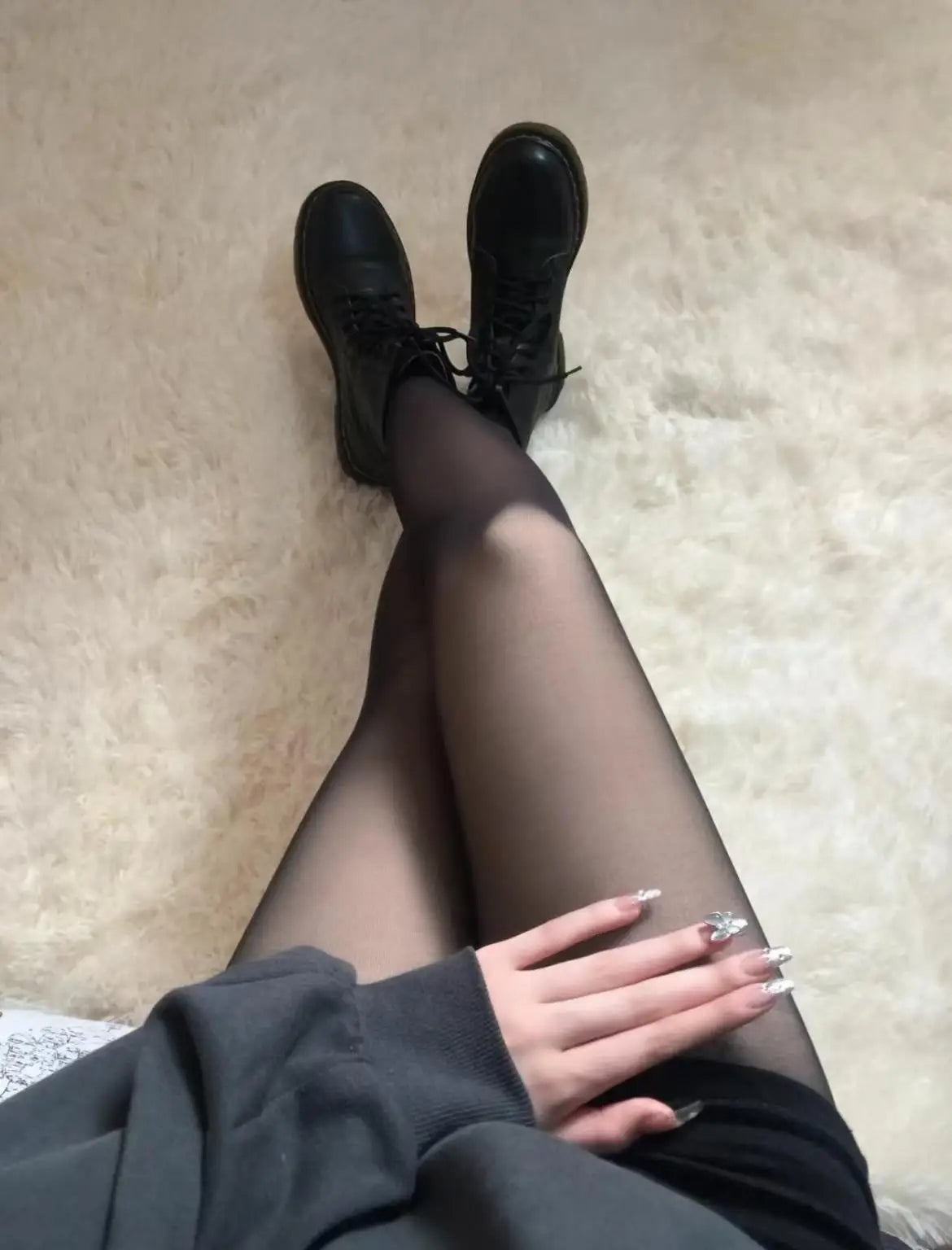 Kvinde iført sorte strømpebukser og sorte støvler, sidder på hvidt tæppe. Hånden med dekorerede negle hviler på låret. Fashion, stil, mode.
